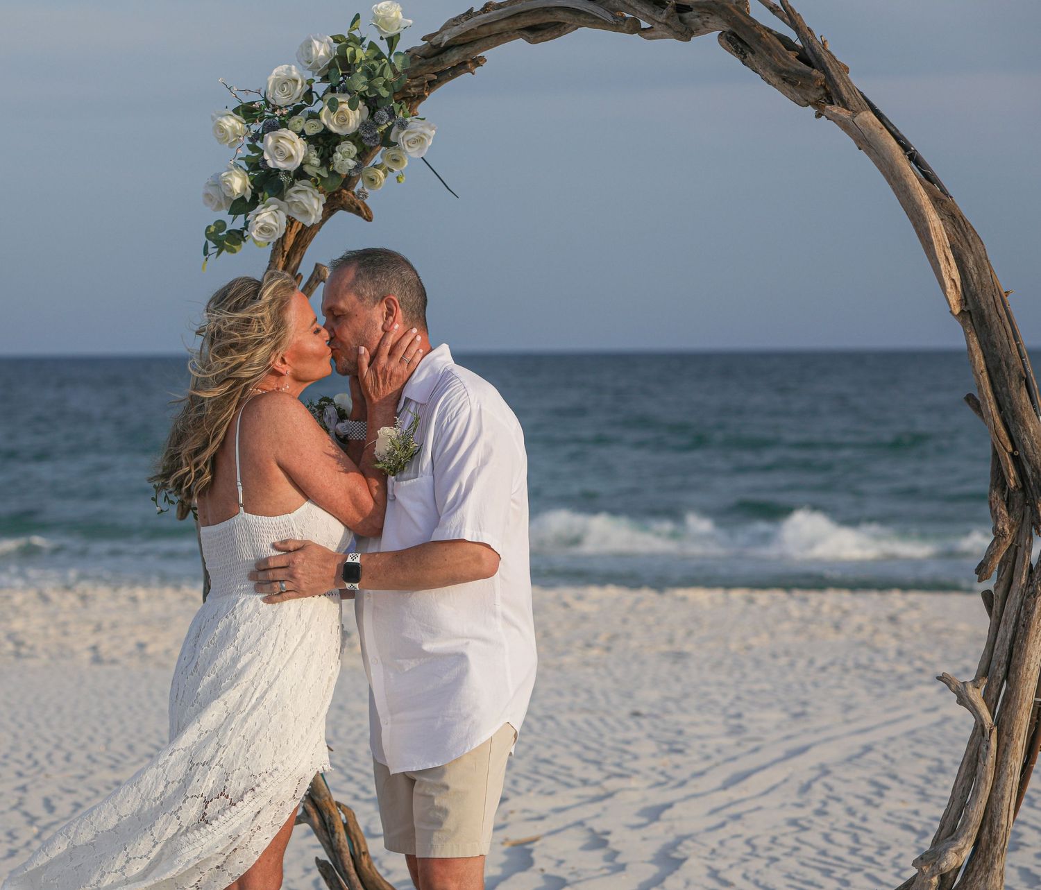A romantic beach wedding moment under a driftwood arch decorated with white flowers.