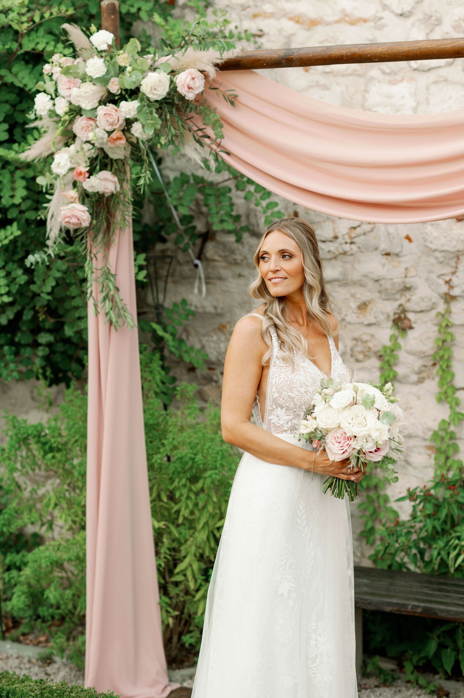 Bride holds pink and white bouquet under romantic floral arch with blush draping.