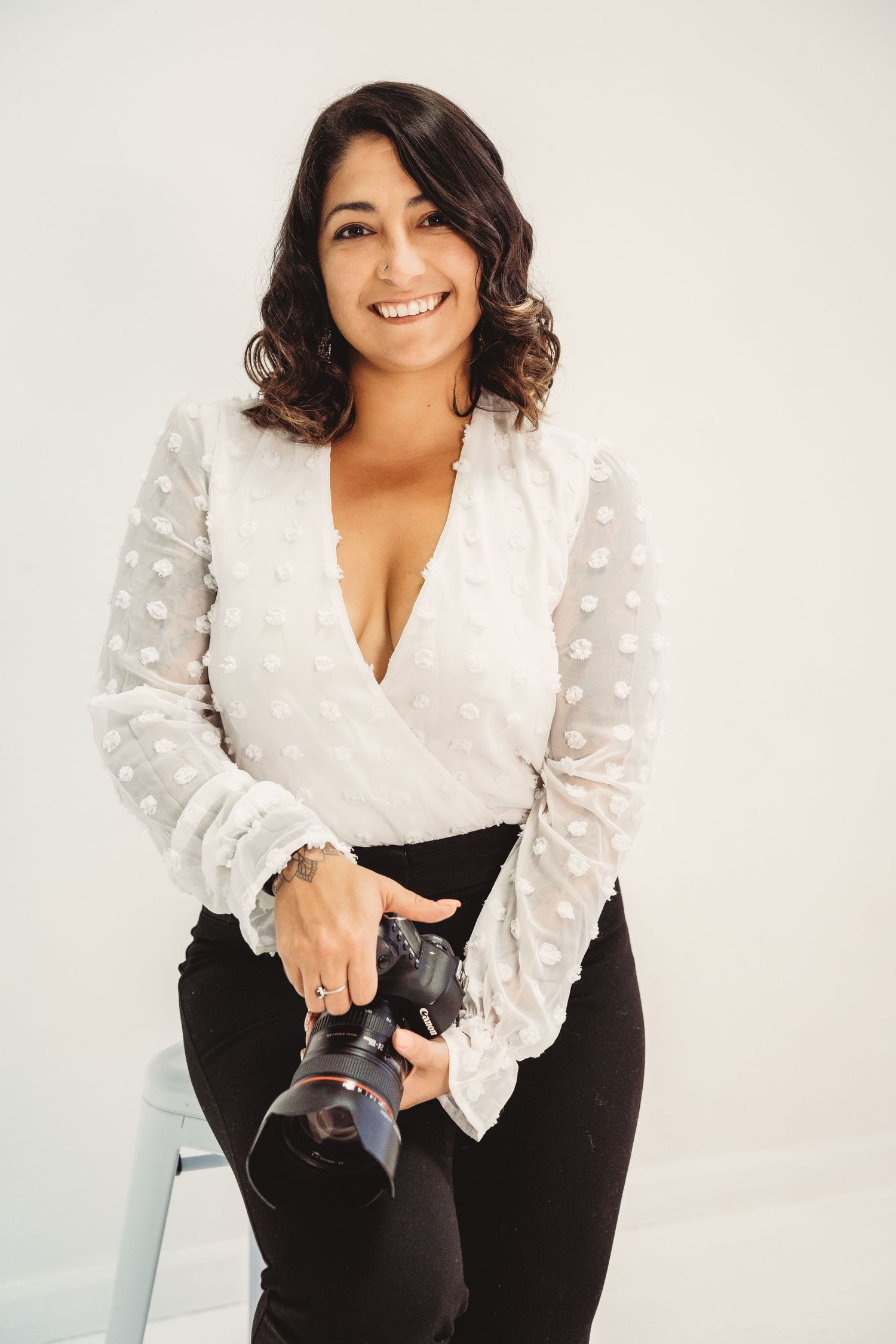 Professional photographer in white blouse and black pants holds camera while posing against white background.