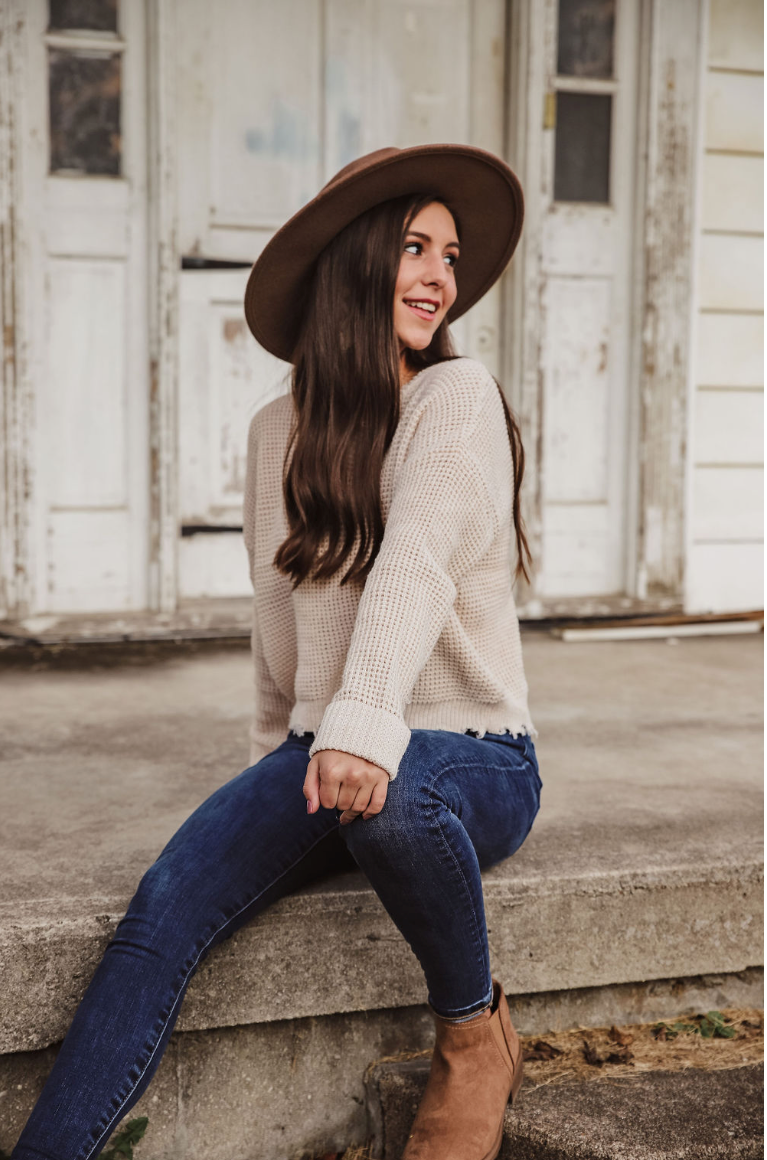 Stylish portrait of a woman in sweater and jeans sitting on steps wearing a wide-brimmed hat.