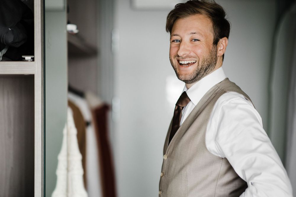 A groom wearing a white dress shirt and beige vest smiles joyfully on his wedding day.