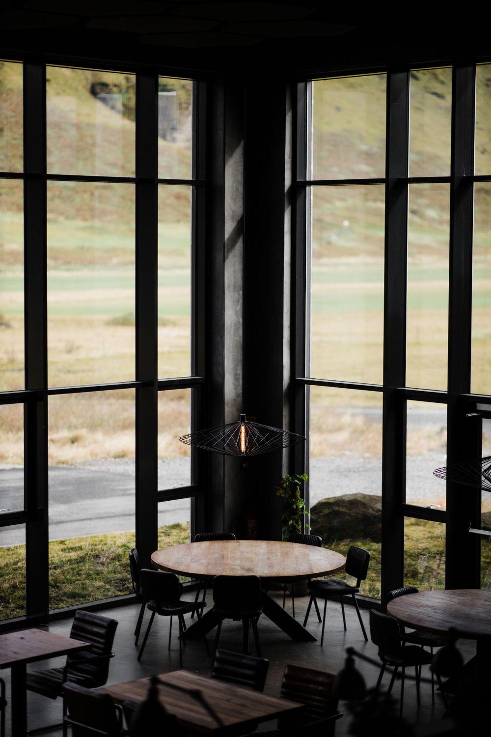 A dimly lit cafe interior with round table and chairs silhouetted against large windows overlooking a scenic outdoor view.