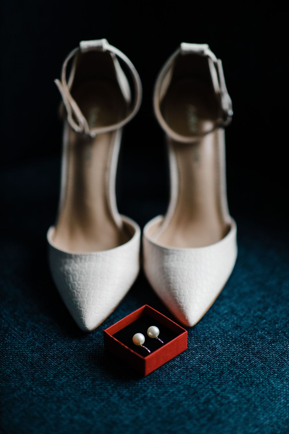 White pointed-toe heels with ankle straps and pearl earrings in red box on blue fabric.