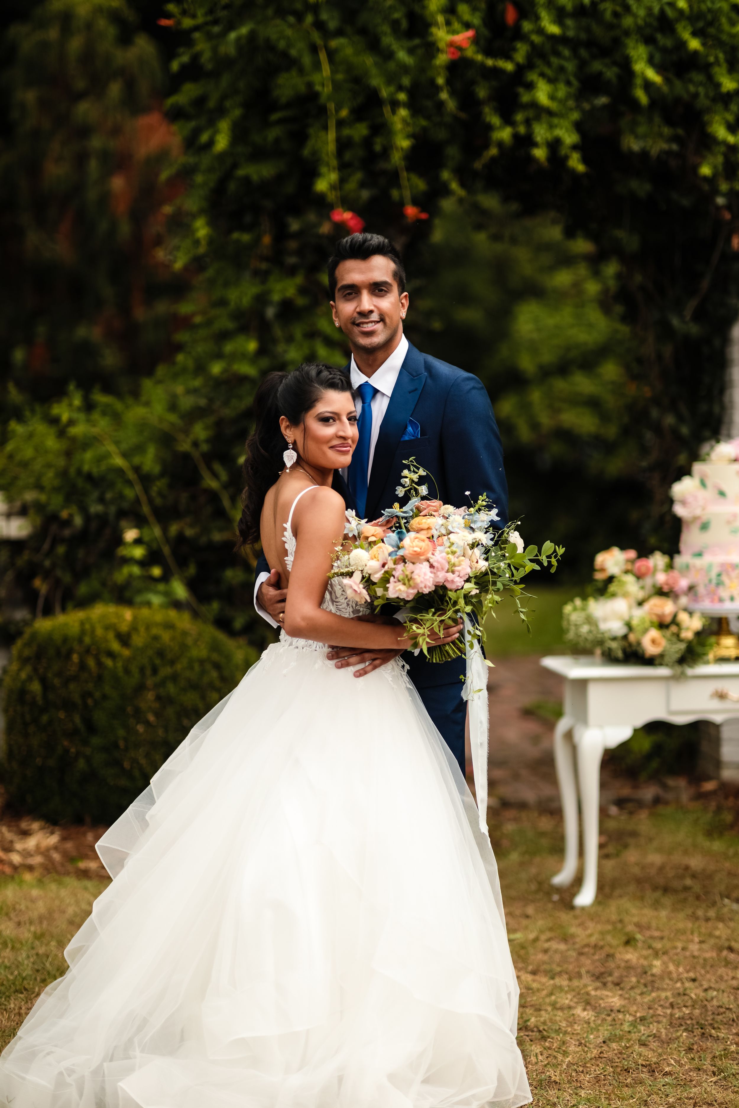 A newlywed couple poses outdoors with a beautiful floral bouquet against a garden backdrop.
