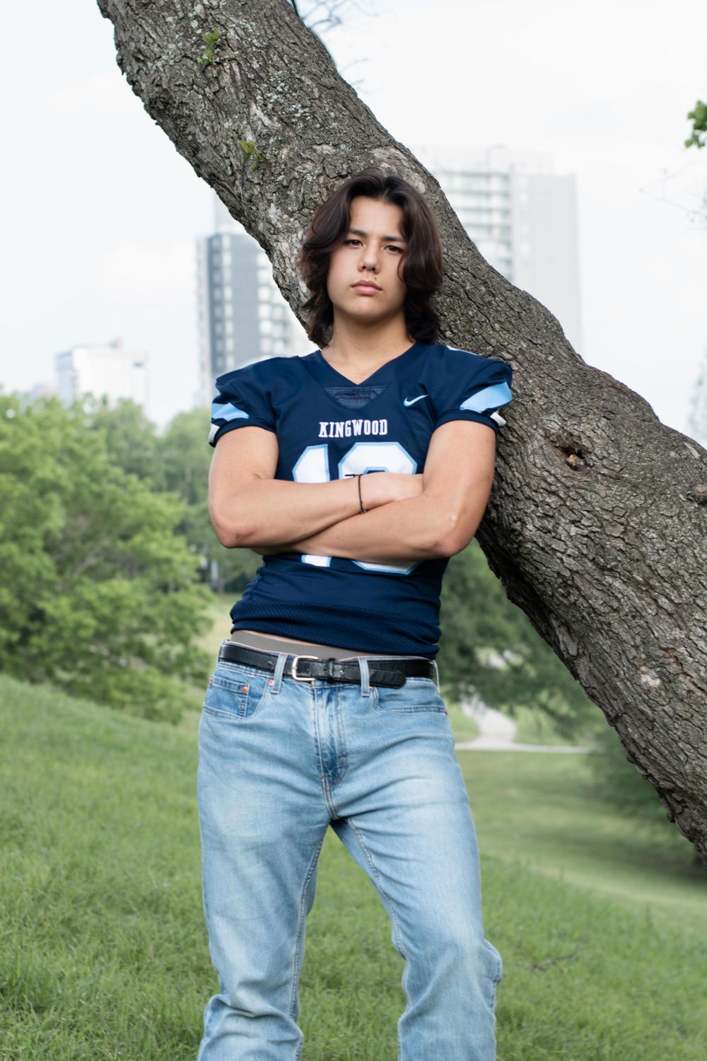 Athletic figure in a navy football jersey and jeans leaning against a large tree trunk in an urban park.