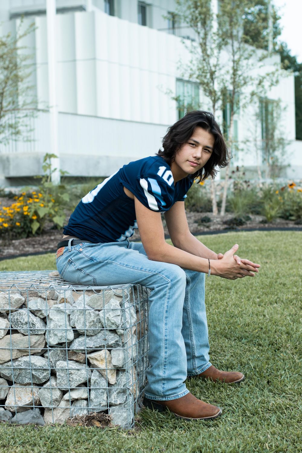 A casual portrait showing someone seated on a stone wall in a blue jersey and jeans on a grassy lawn.