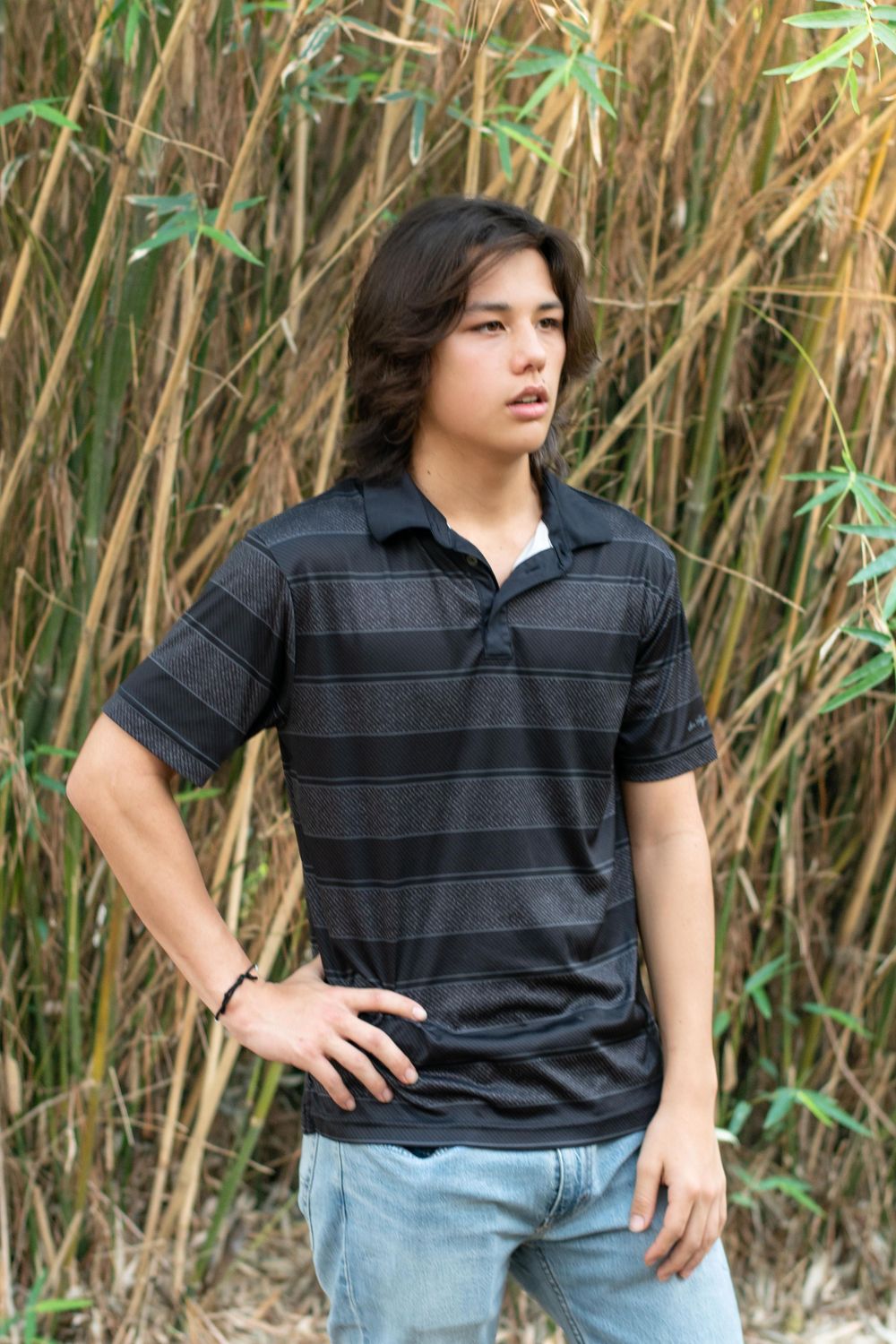 Casual portrait against bamboo background showing person in striped polo shirt and jeans with long dark hair.