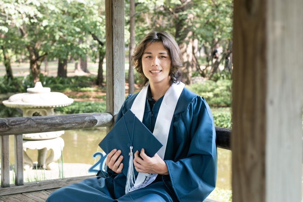 Graduate seated in traditional blue academic regalia near a decorative pond.