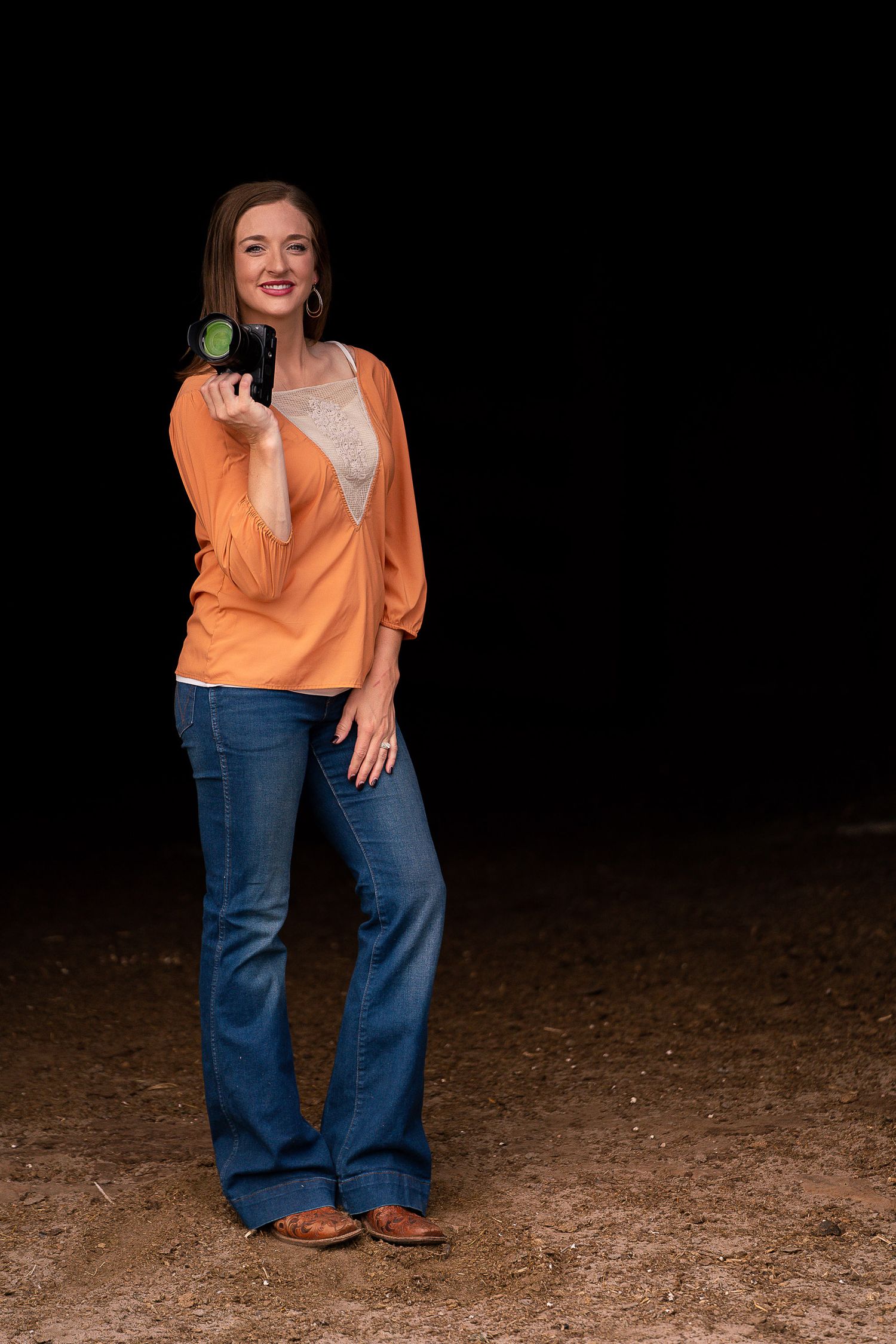 Someone in a bright orange top and flared jeans poses outdoors on a dirt ground at night.