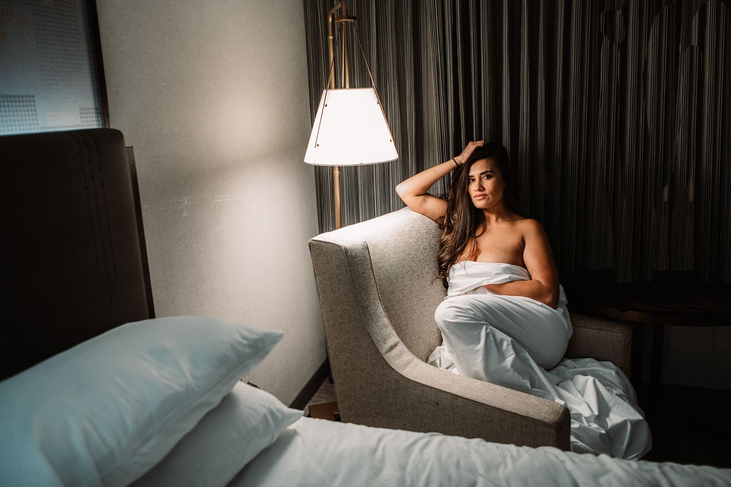 Intimate bedroom portrait with soft hotel lighting and white bedding.