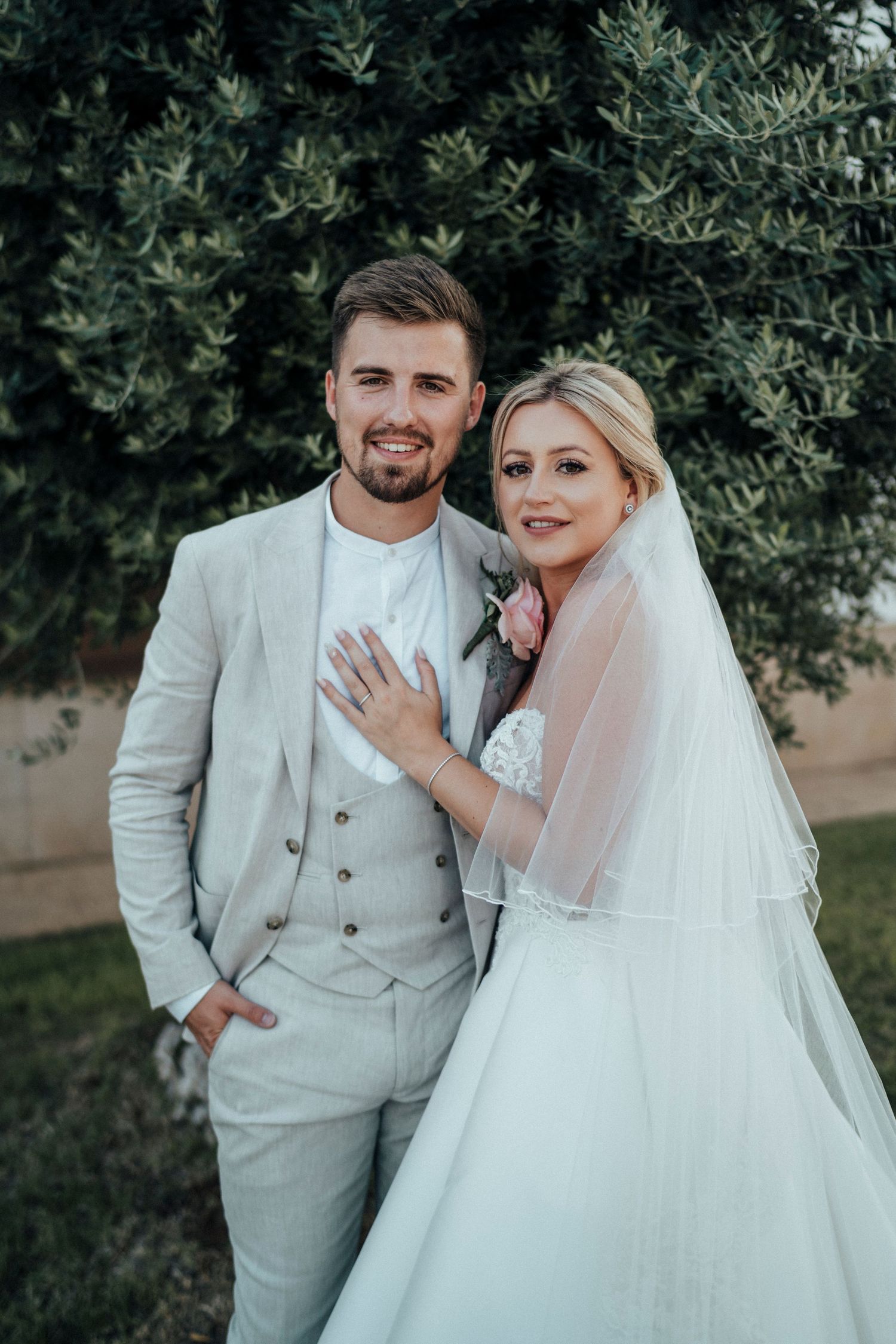 A couple in elegant wedding attire poses against a lush green hedge backdrop.