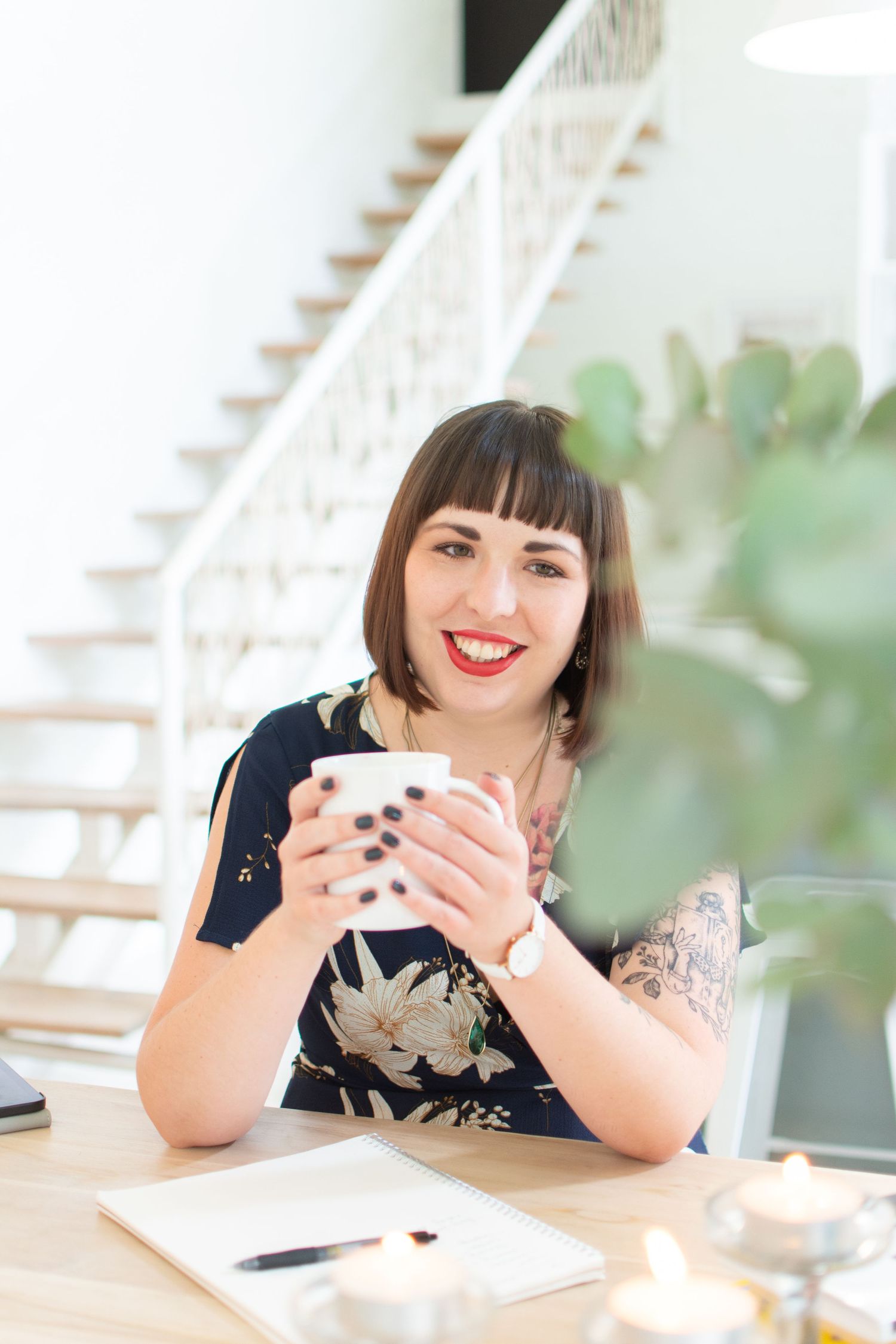 Someone enjoying coffee at a desk with a notebook and plants in the background.