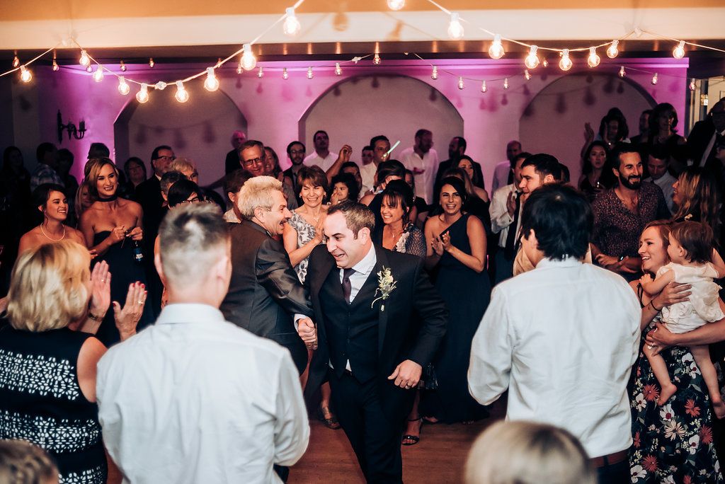 Crowd of wedding guests dance and celebrate on a reception dance floor with purple accent lighting.