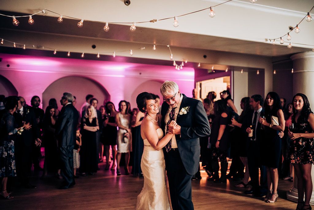 A couple dances together in an elegant ballroom with pink accent lighting and string lights overhead.