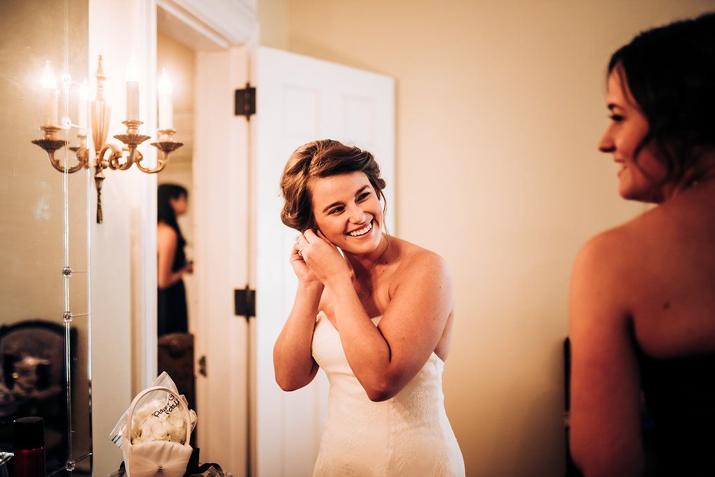 A woman in a white dress putting on earrings in front of a mirror.