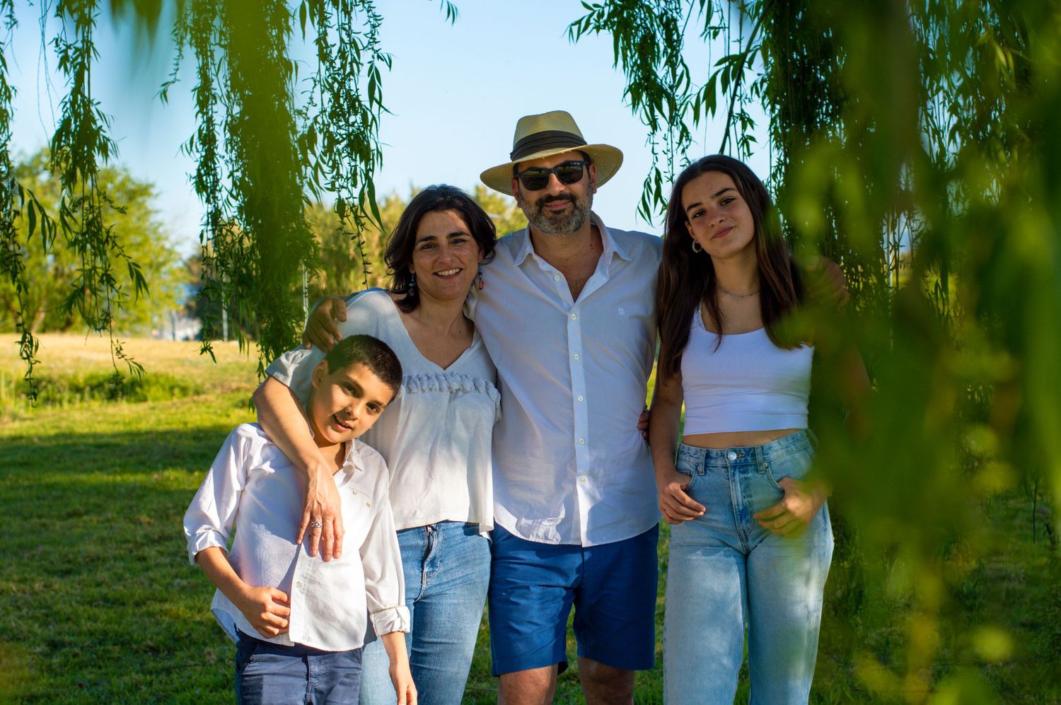 A family of four poses together under a weeping willow tree on a sunny day.