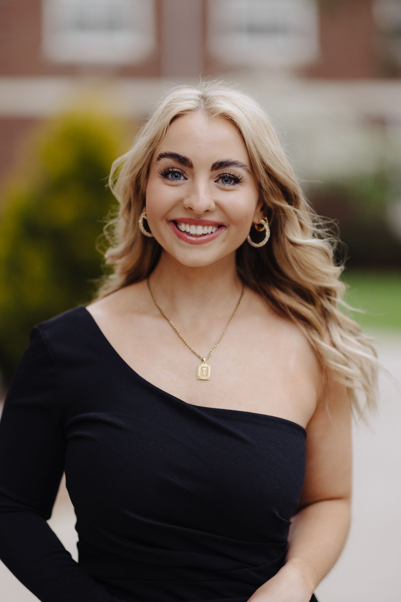 Woman in elegant black asymmetrical top smiles warmly in outdoor portrait.