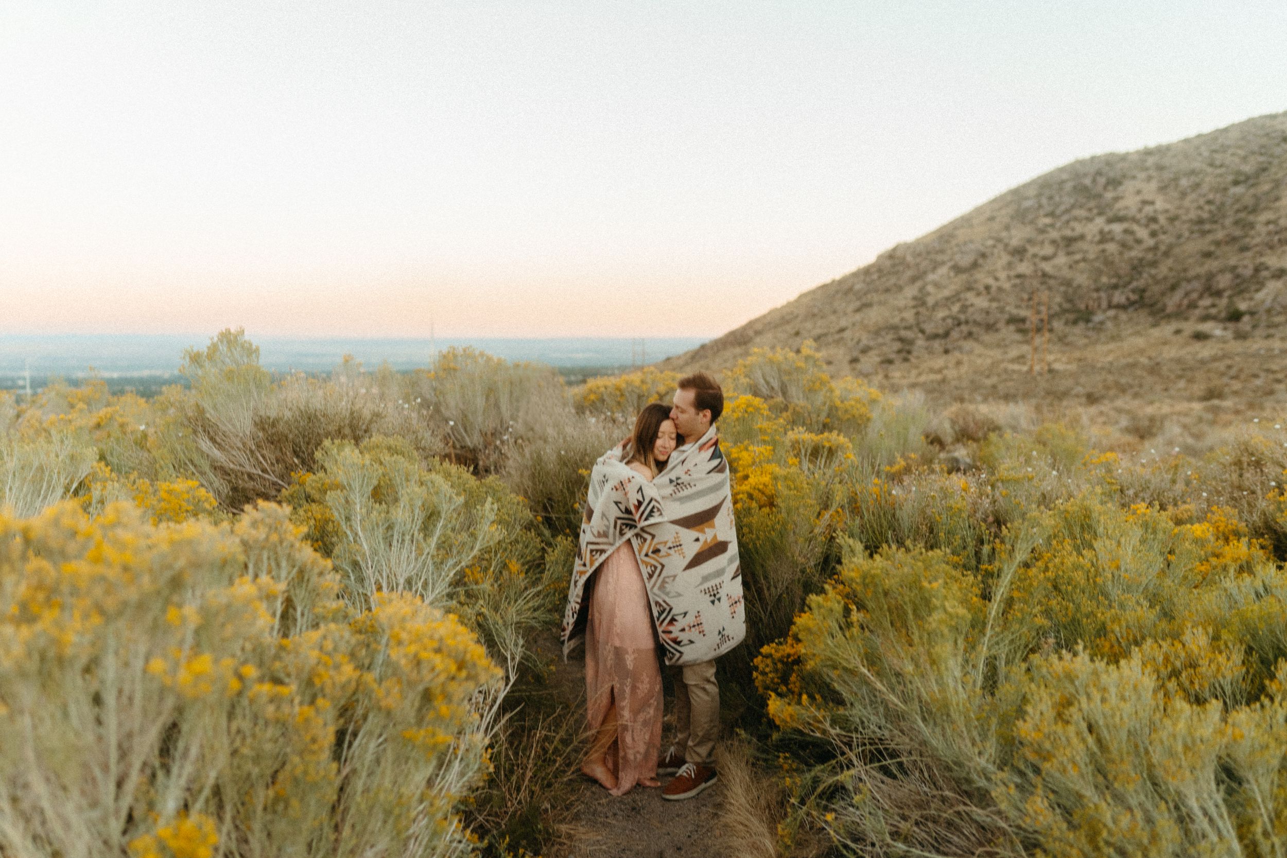 A couple embraces in a desert landscape with yellow sagebrush and mountains during golden hour.