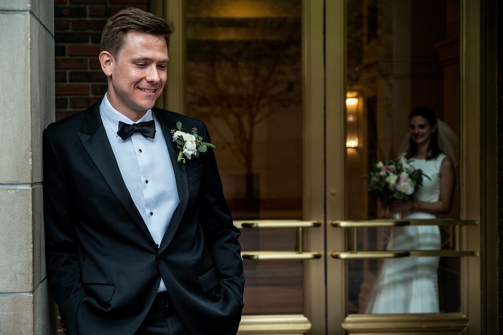 A groom in classic tuxedo poses while bride is reflected in glass doors behind him.