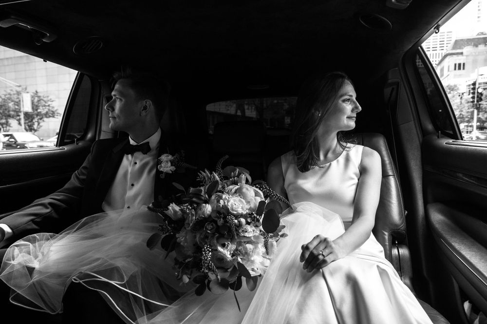 A black and white photo of newlyweds sitting in their wedding car with bouquet.
