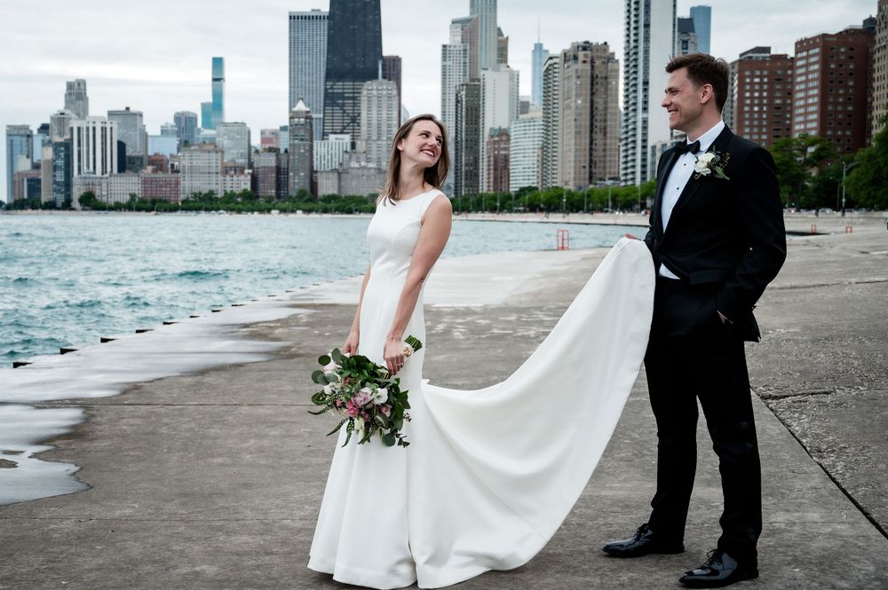 Wedding couple poses dramatically against Chicago skyline with flowing dress and Lake Michigan waves.