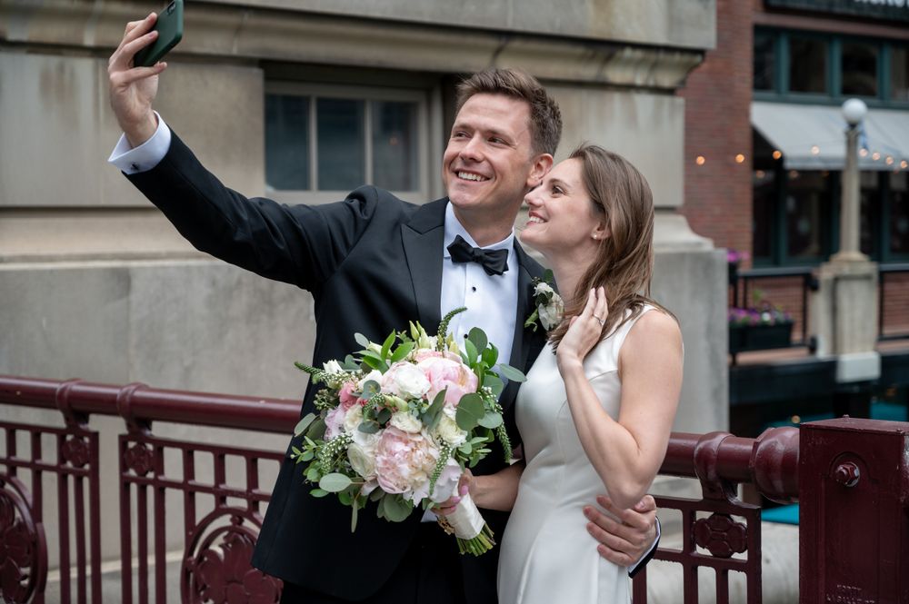 Wedding couple takes selfie on historic bridge while holding pink and white floral bouquet.
