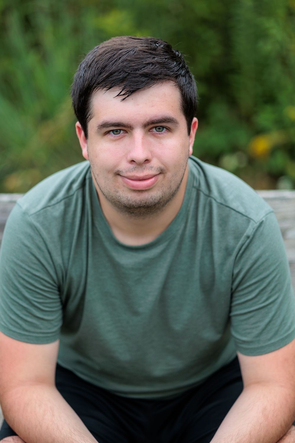A person in a sage green t-shirt sits outdoors with a gentle smile against a blurred natural background.