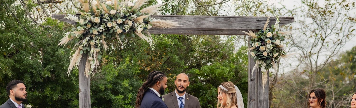Wedding ceremony under rustic wooden arch decorated with white floral arrangements and greenery.
