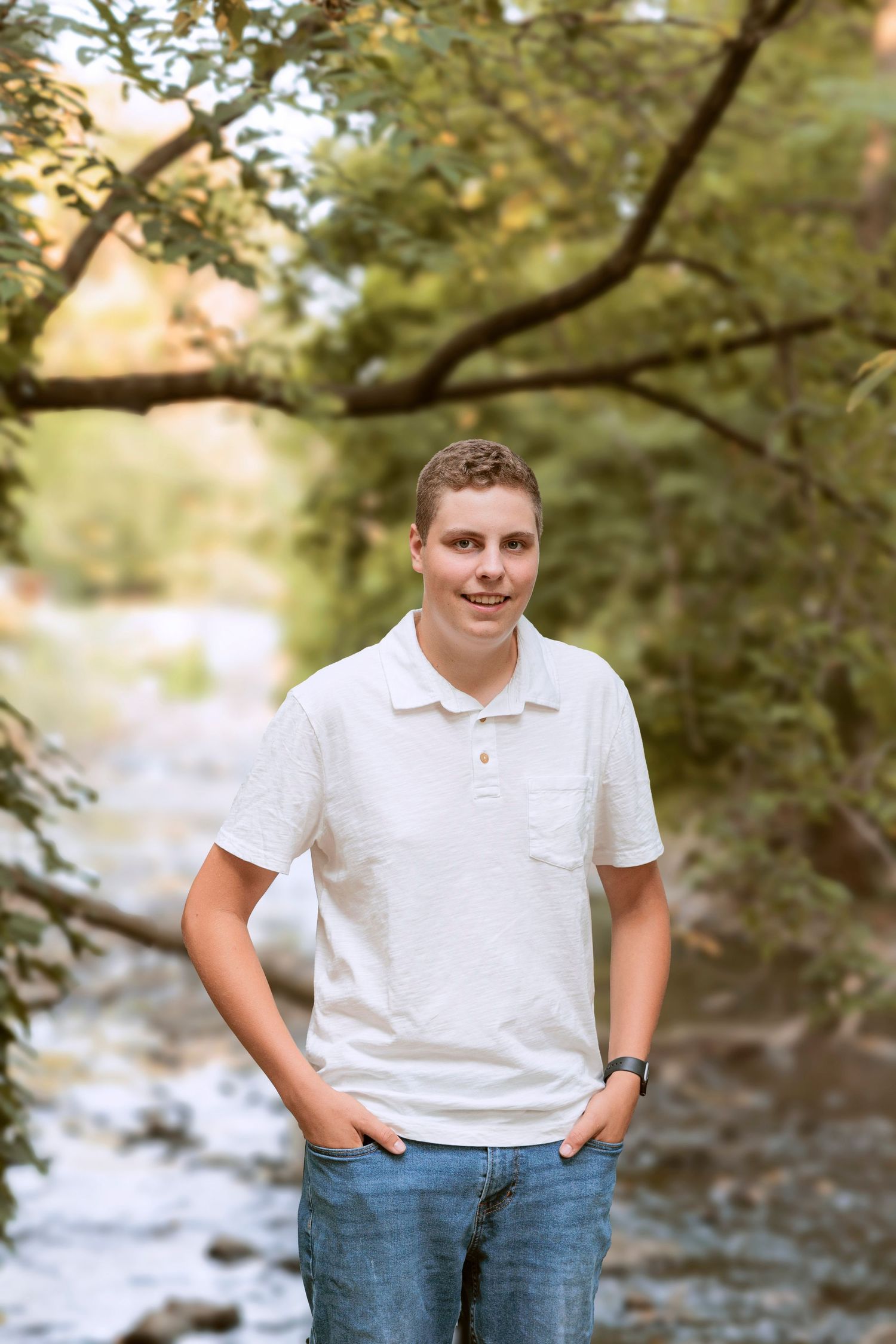 Portrait in white polo shirt by stream with trees in background.