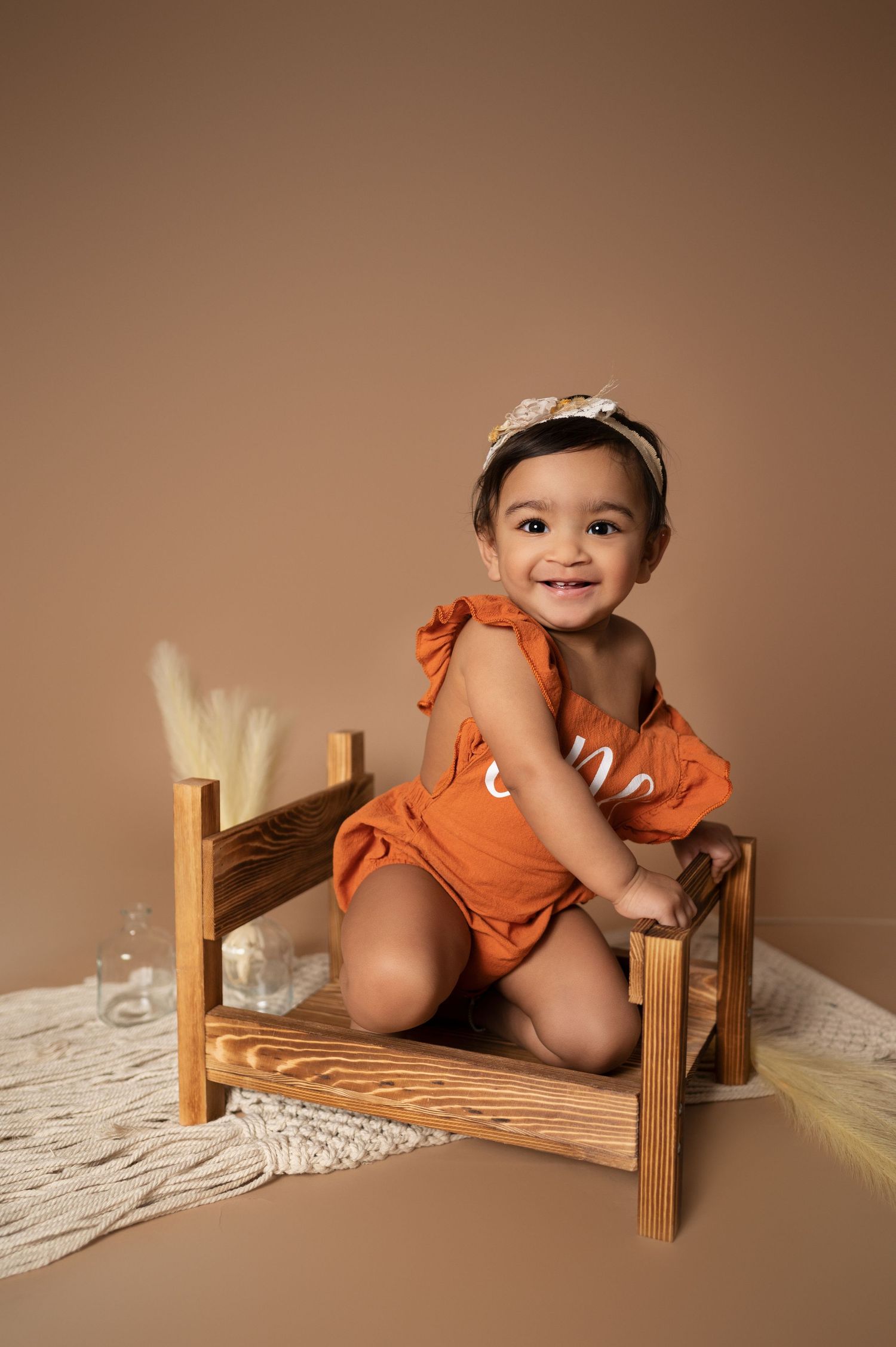 Baby in orange outfit sits on wooden bench against brown backdrop during photoshoot.