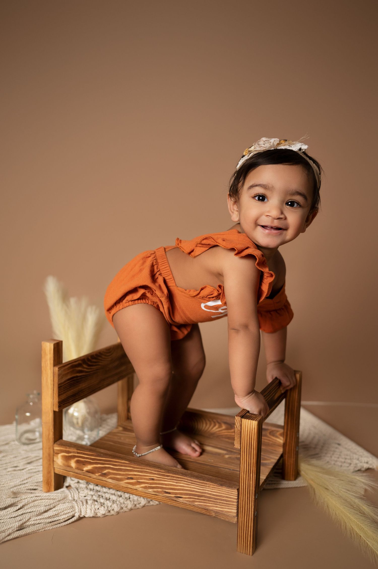Baby wearing orange romper poses on wooden bench during professional photoshoot.