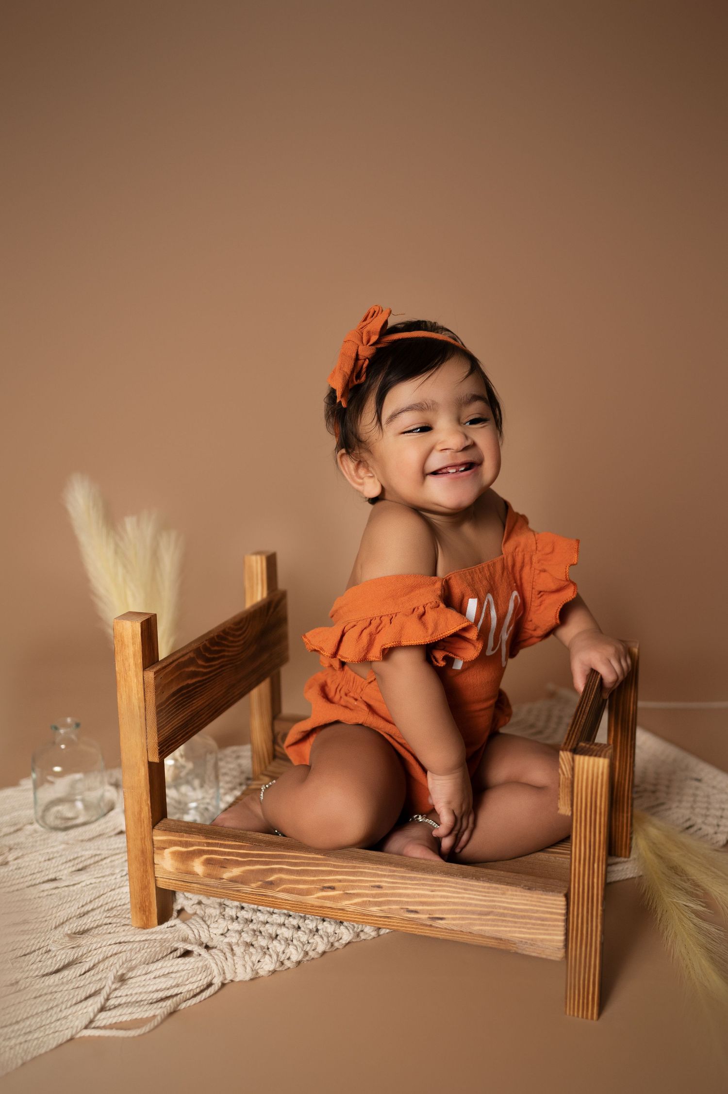 Baby in ruffled orange outfit smiles while sitting on wooden bench with macrame decor.