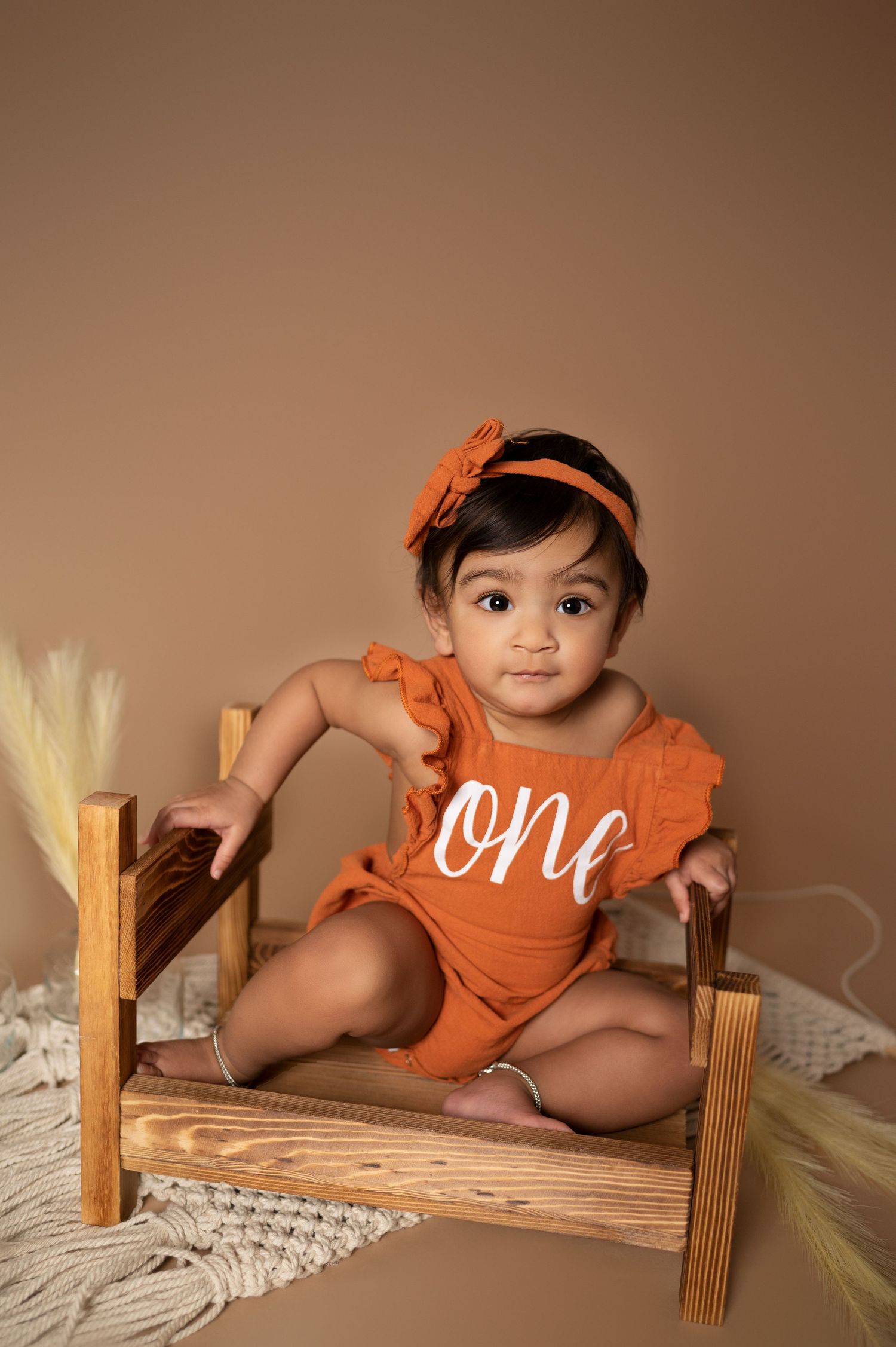 Baby in orange 'one' romper poses on wooden bench for first birthday photoshoot.