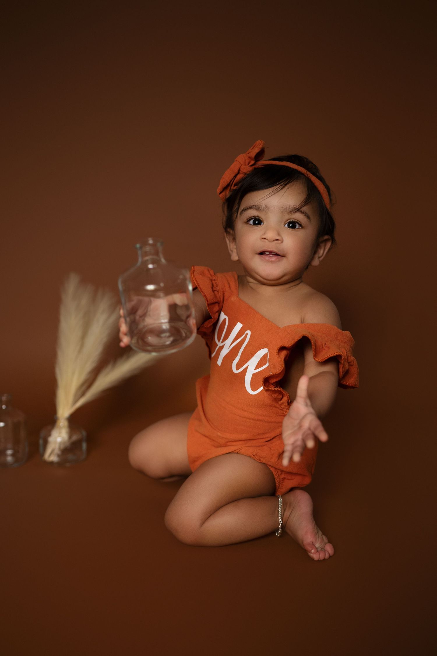 Baby in orange outfit with headband sits on brown backdrop with wheat grass decor.