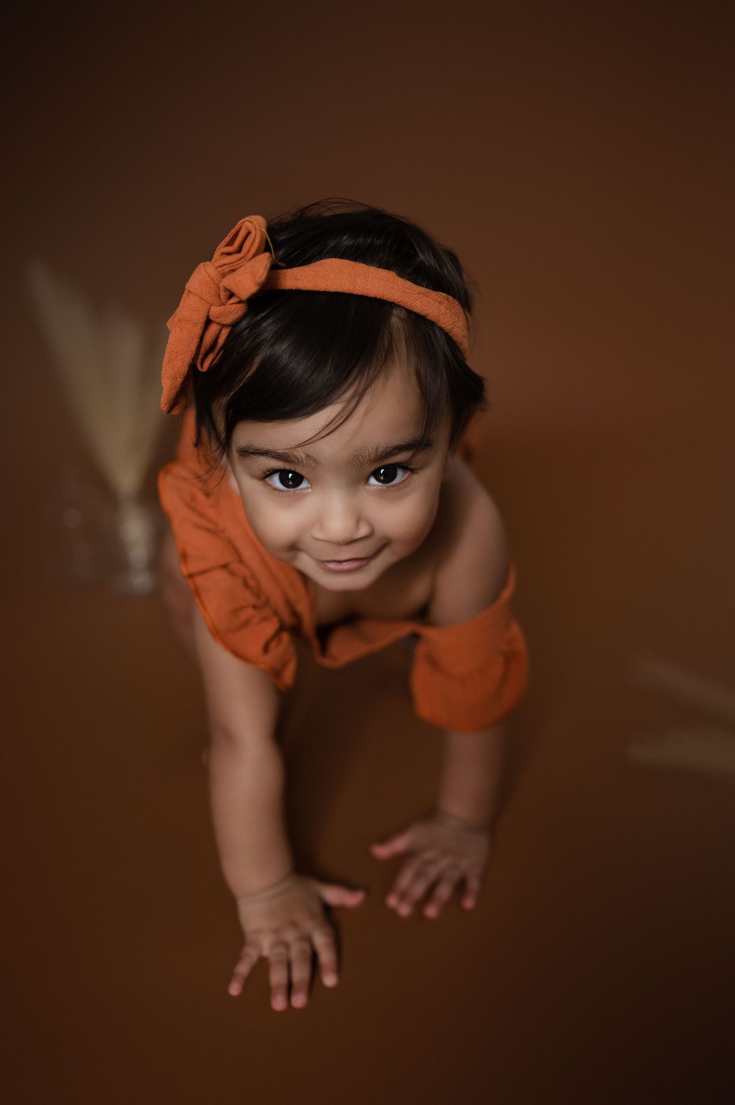 Baby in orange outfit crawls on brown backdrop during professional photoshoot.