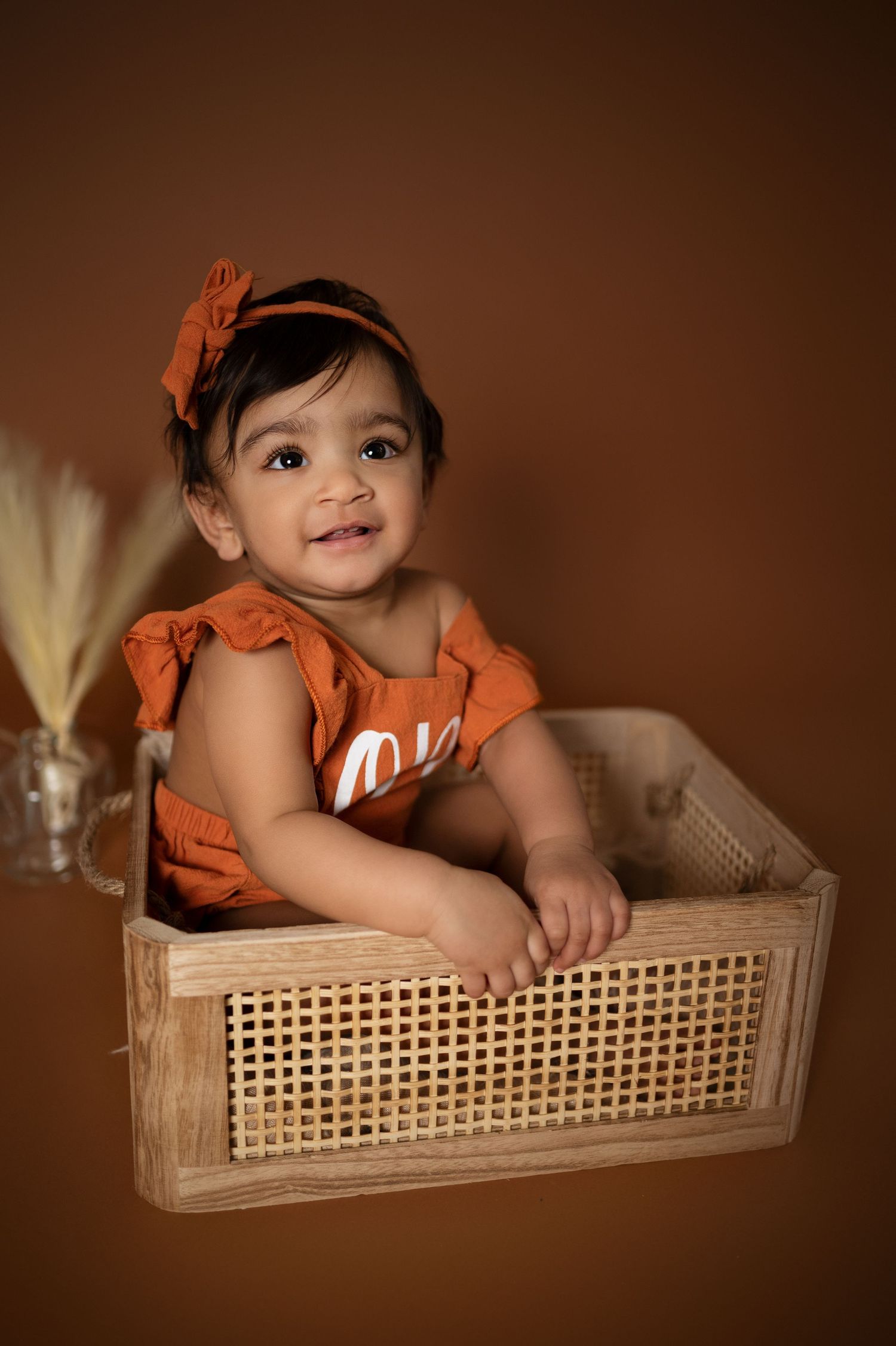 Baby in orange romper sits in wooden cane box during studio photoshoot.