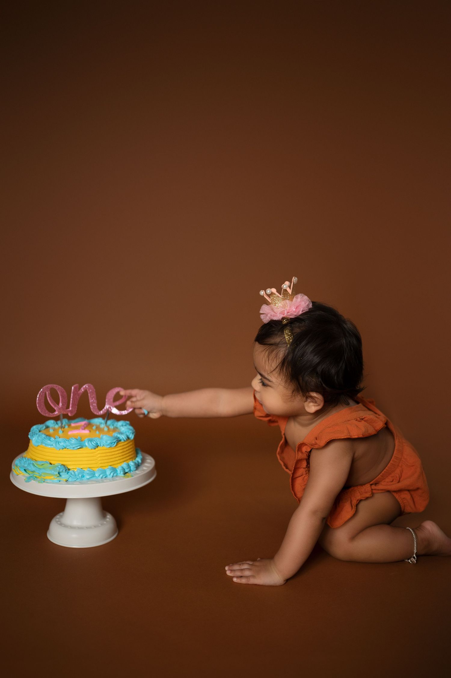 Baby in orange outfit leans toward blue and yellow birthday cake during photoshoot.