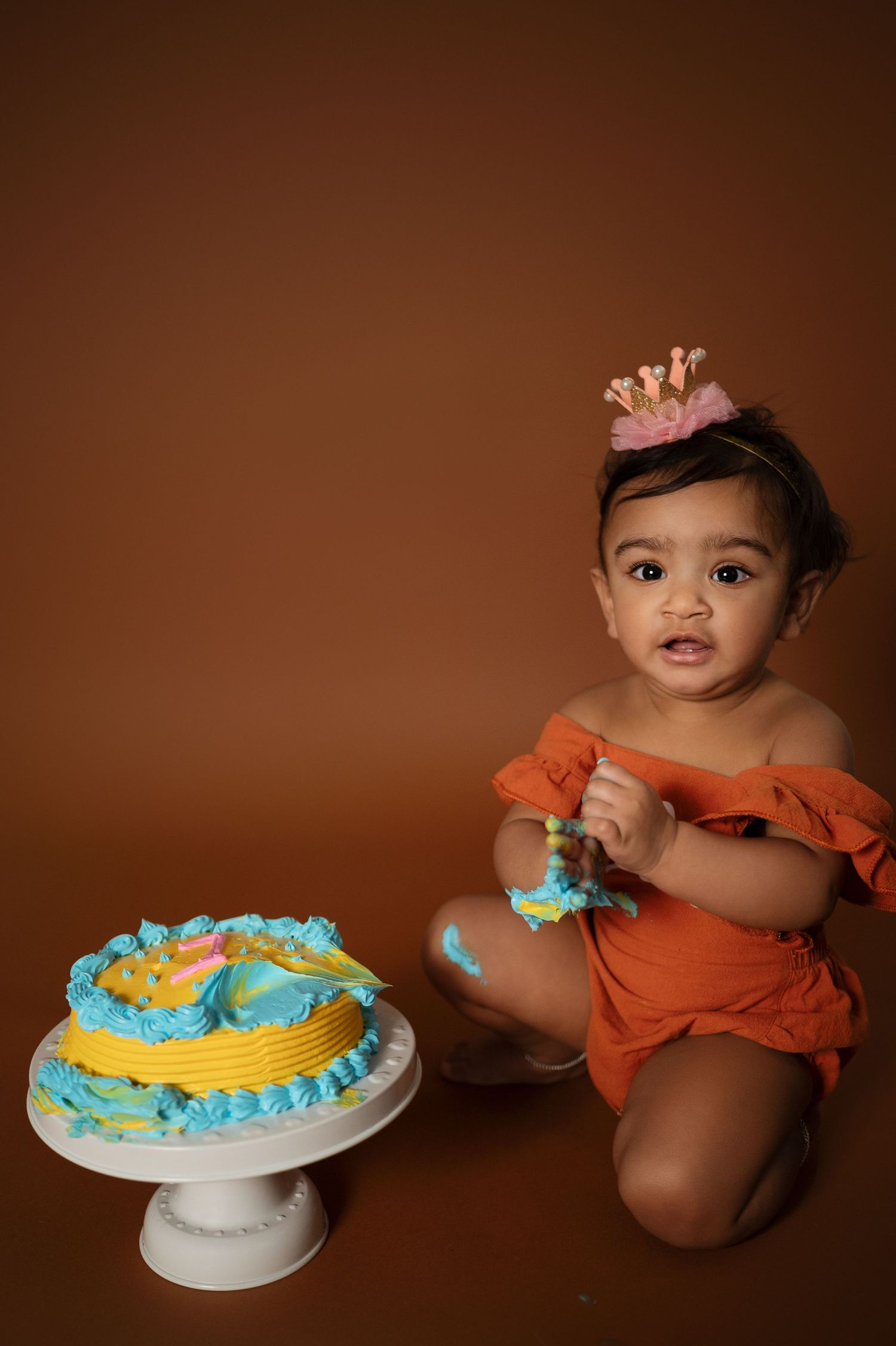 Baby explores blue and yellow birthday cake while wearing orange outfit during cake smash session.
