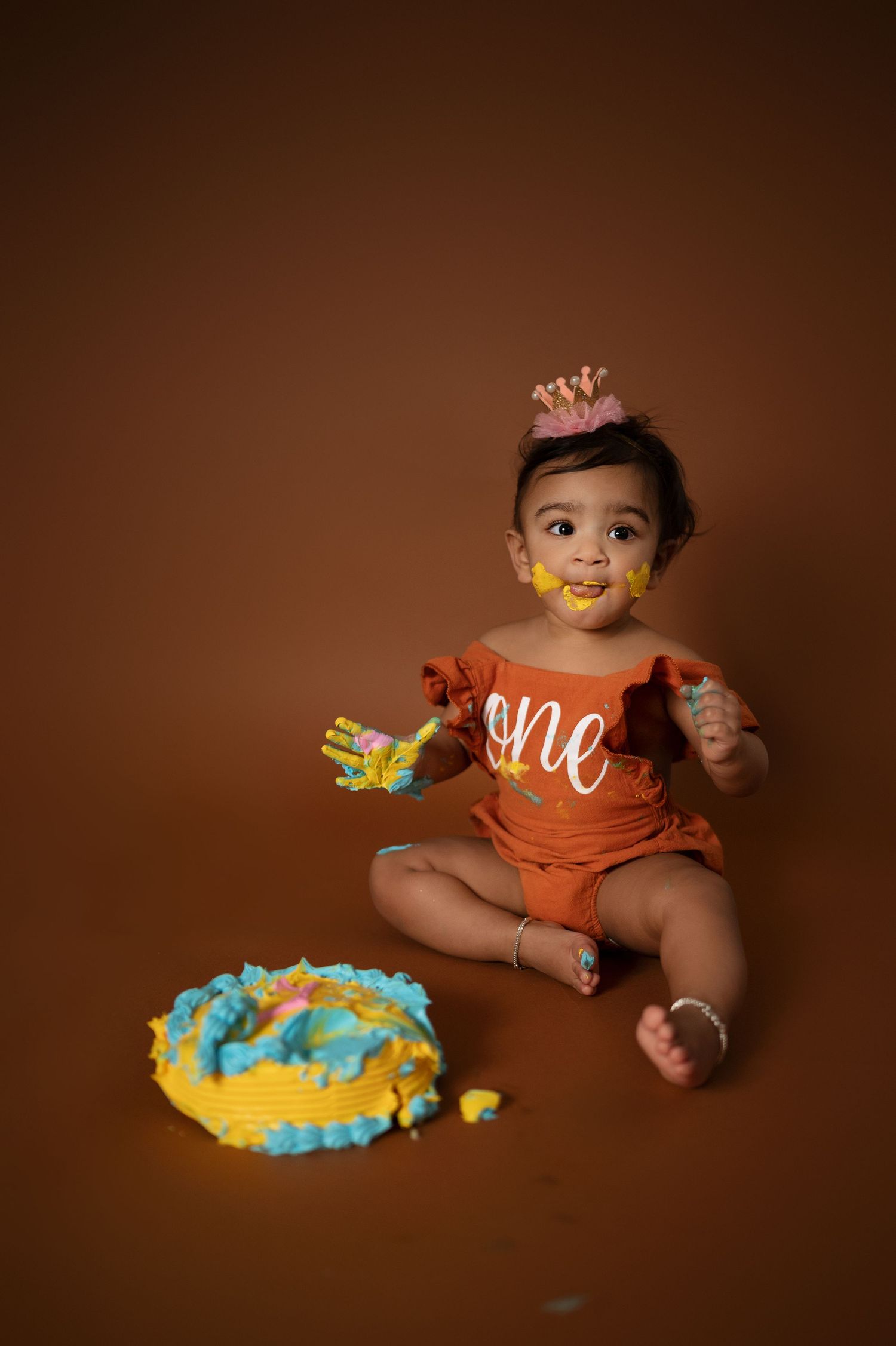 Baby wearing orange outfit and crown smashes colorful birthday cake during first birthday photoshoot.