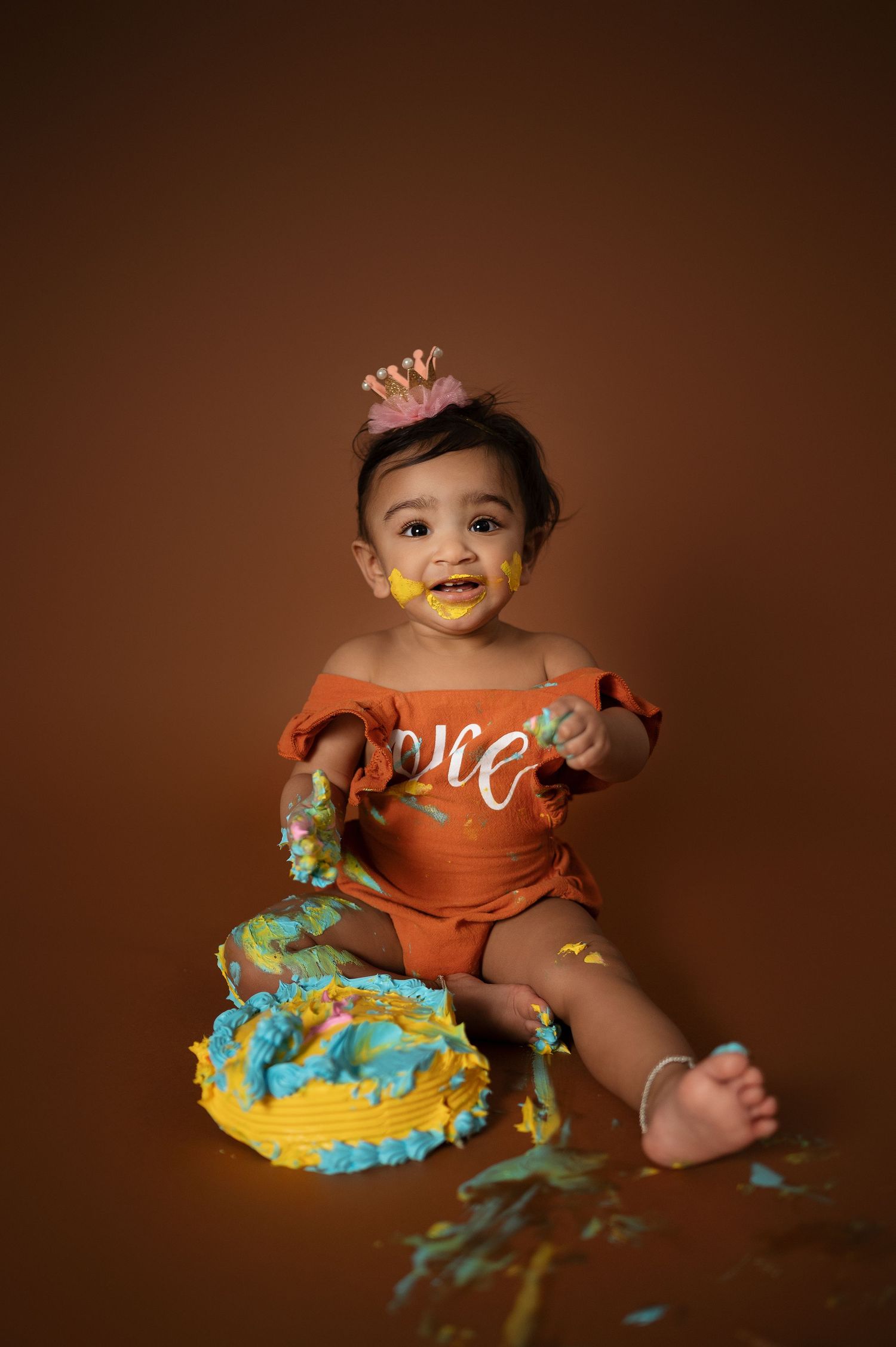 Baby in orange romper with crown plays with smashed blue and yellow cake.