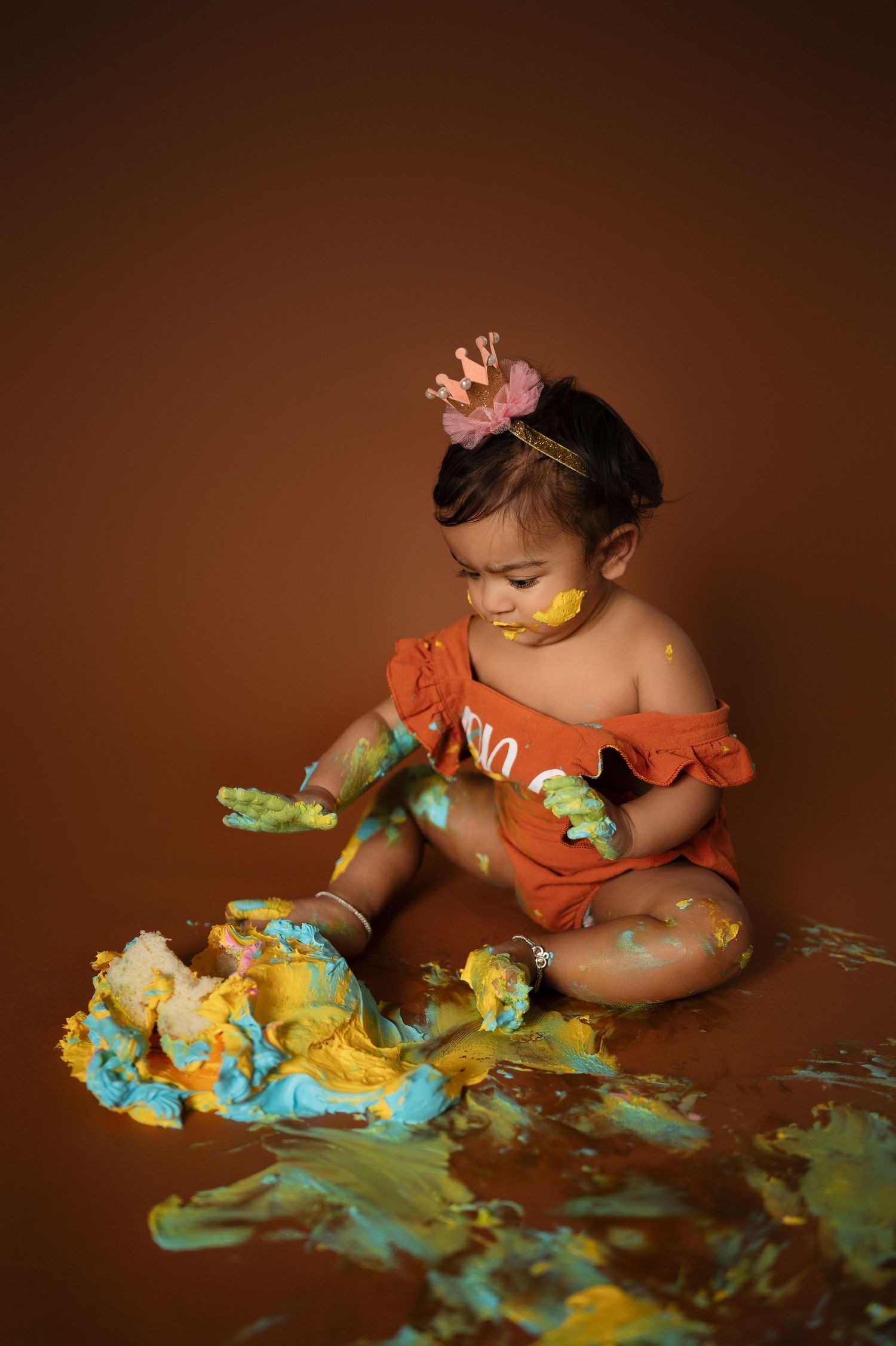Baby in orange outfit plays with smashed blue and yellow frosting on brown background.