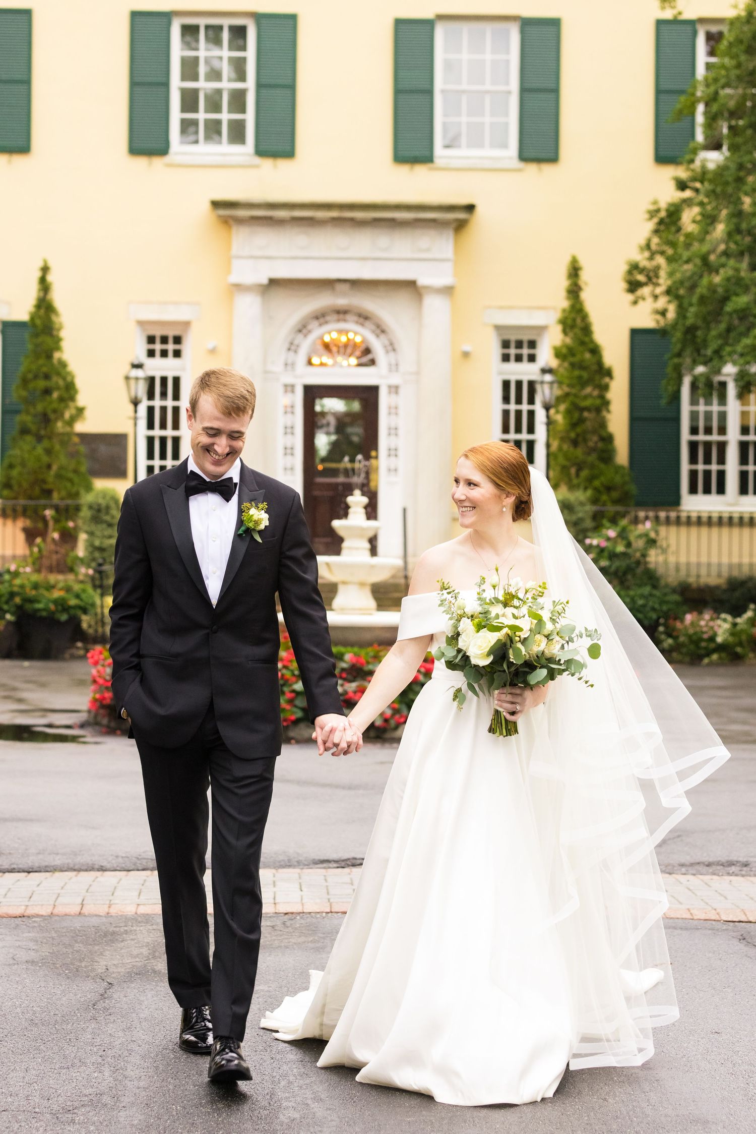 An elegant couple walking outside a yellow colonial mansion with green shutters.
