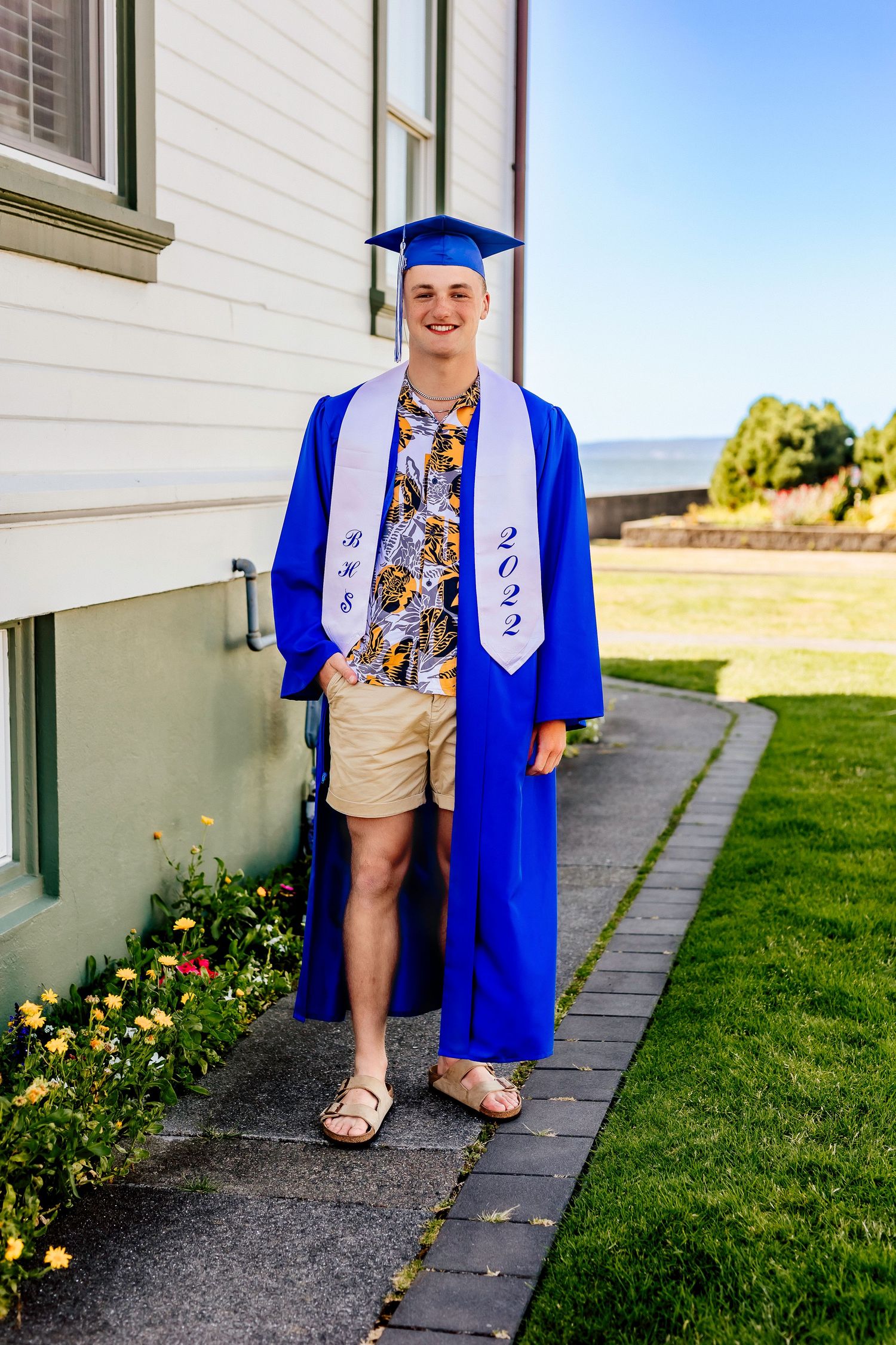 Graduate in blue cap and gown poses outside white building with landscaped garden.