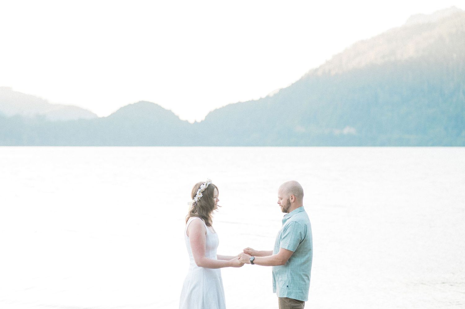 A couple stands together on a lake shoreline with mountains in the background during their elopement at Lake Crescent