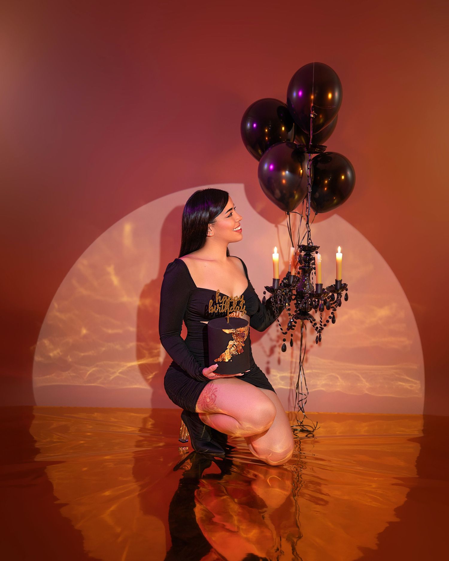 Woman in black dress posing with dark balloons and candelabra against sunset backdrop.