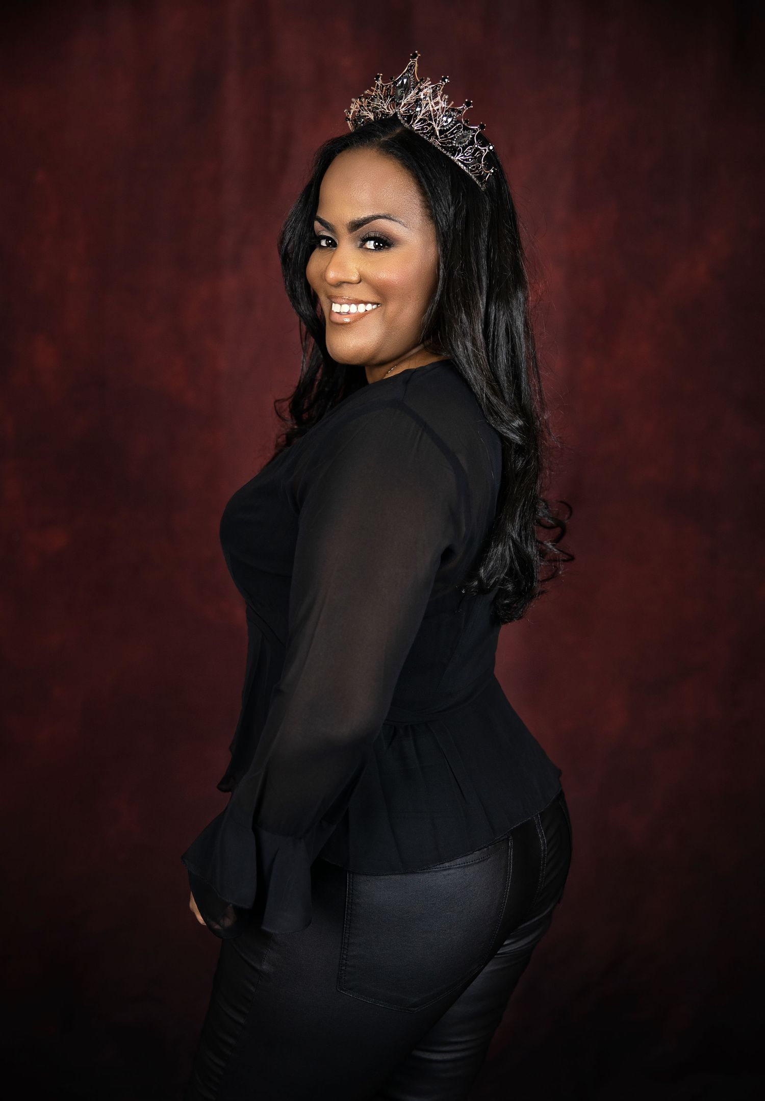 Glamorous portrait against burgundy backdrop featuring an elegant black outfit and crown.