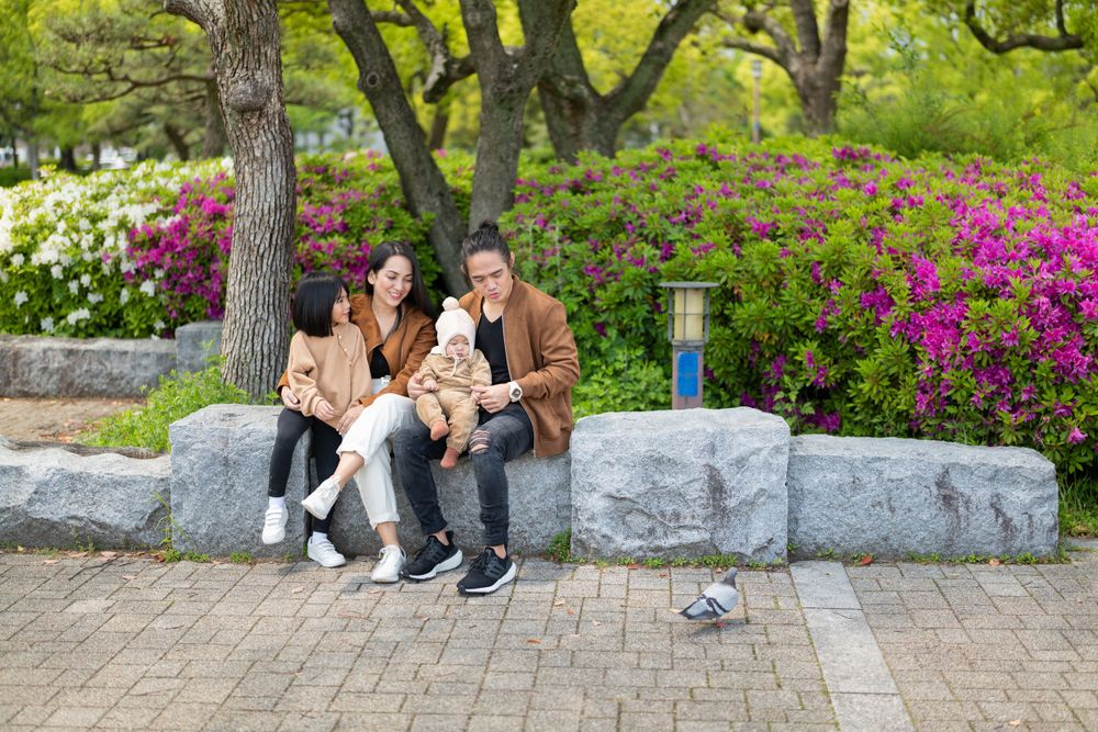 Family of four relaxing on a stone bench in a vibrant park with pink and white flowering bushes in Japan.
