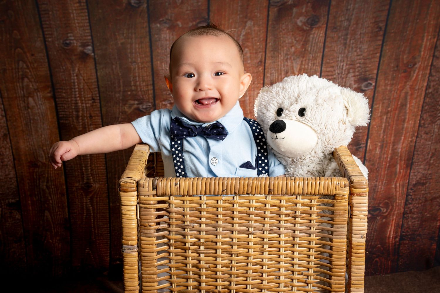 A baby in a blue bowtie sits in a wicker basket with a plush teddy bear. Scotch Plains Photographer, portrait photography.