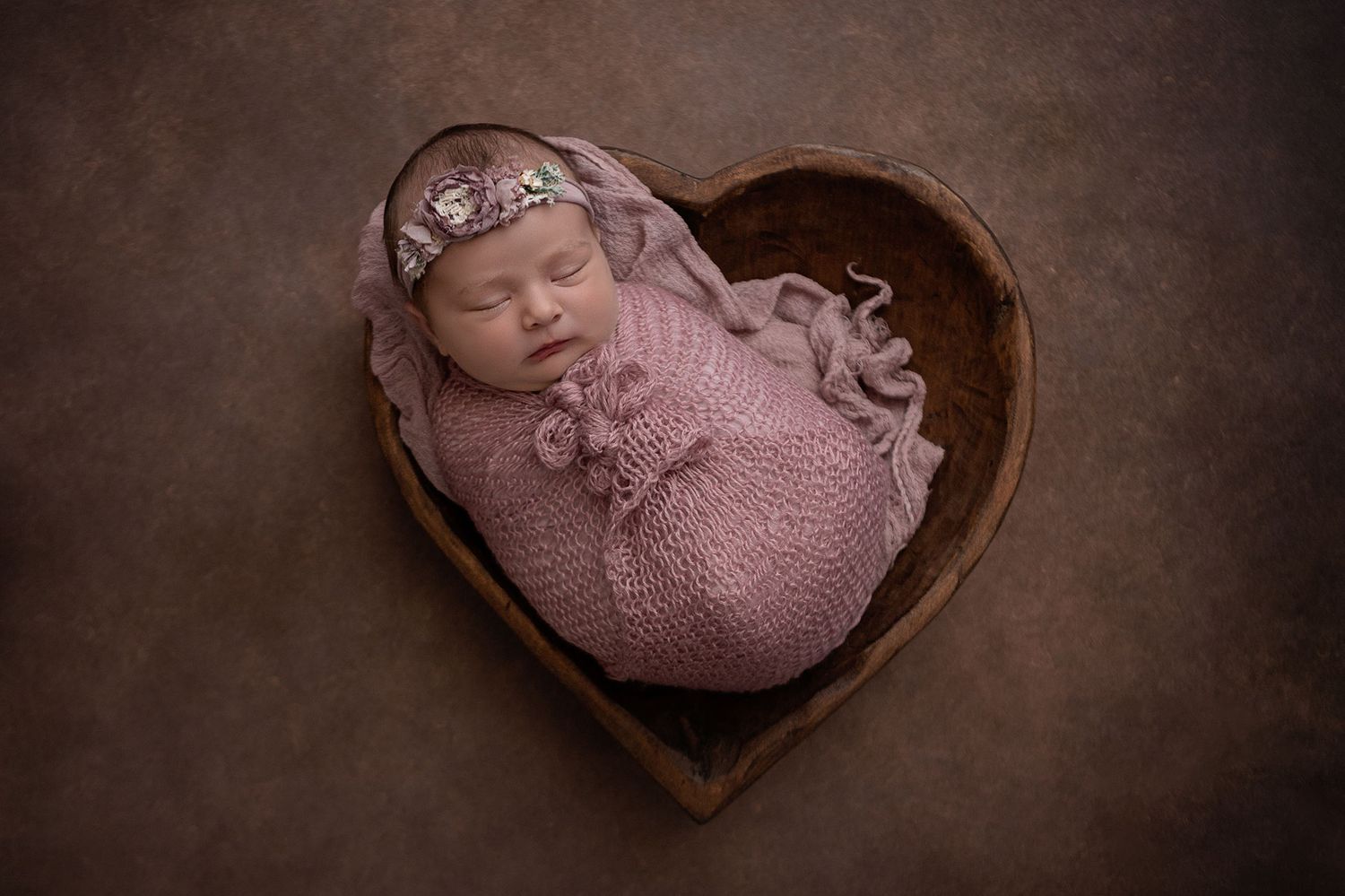 Newborn baby girl wrapped in pink wrap laying in a wood heart bowl in Prosper, Texas.