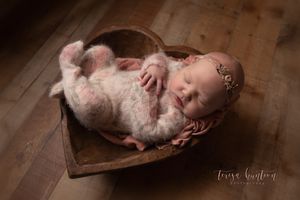 Newborn baby girl in floral onesie wearing a pink floral headband laying in a wood heart bowl in Dallas, Texas.